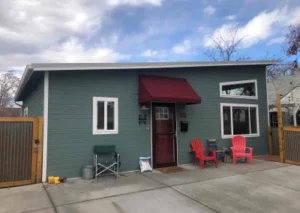 A modern green house with a red awning, featuring white windows and two red chairs on the porch. A driveway leads up to the entrance